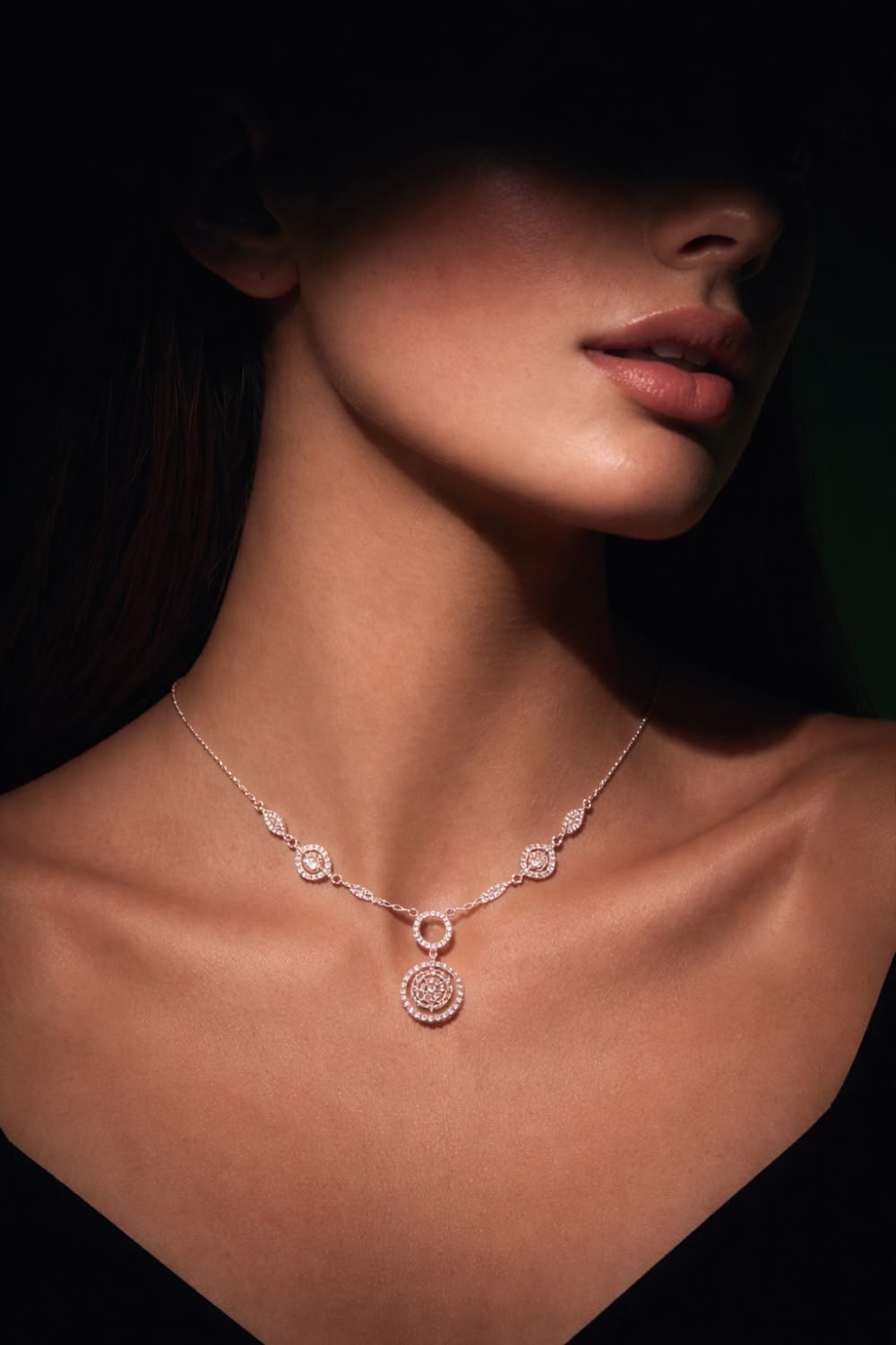 A woman wearing a diamond necklace with a circular pavé pendant, lit from one side against a dark ground.